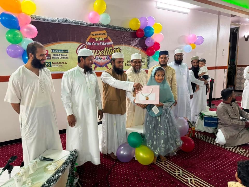 A young girl in a headscarf and dress receives a gift box from men in traditional Islamic attire on a red-carpeted stage decorated with colorful balloons and a banner for the International Kids Qur'an Tilawah Competition.