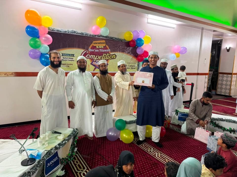 A man in a navy robe holds a certificate presented by a group of men in white robes and caps, standing in front of a balloon-decorated banner reading "International Kids Qur'an Tilawah Competition" inside a mosque hall with a red patterned carpet.