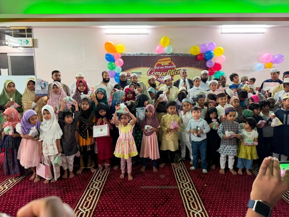 A large group of children in traditional and formal clothing stand together holding small gift bags in a mosque hall decorated with colorful balloons and a banner reading "International Kids Qur'an Tilawah Competition," with several adults, including men wearing kufis, standing among them.