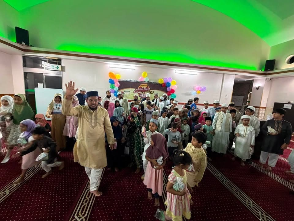 A man in traditional attire raises his hand while leading a large group of children and families, many wearing prayer caps and headscarves, gathered in a hall decorated with colorful balloons and a "Qur'an Tilawat Competition" banner.