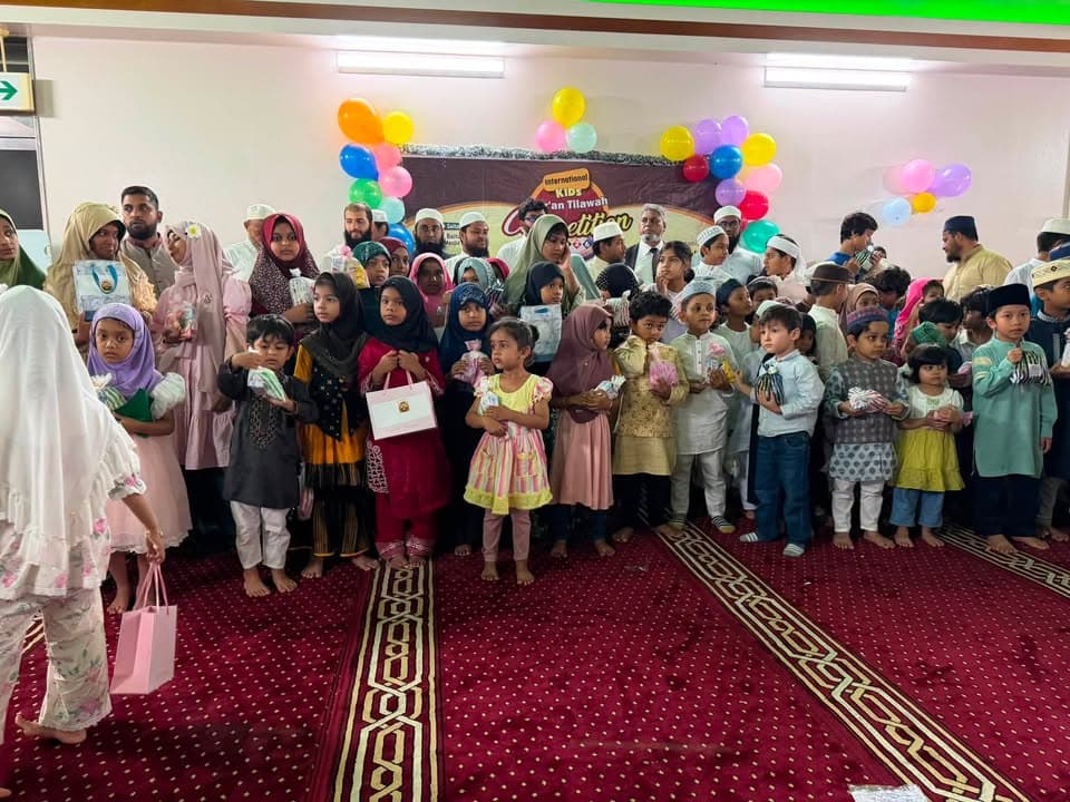 A large group of children and adults stand together on a red carpeted prayer hall, with many children holding certificates and small gift bags, under a wall decorated with colorful balloons and a banner reading "International Kids Qur'an Tilawah Competition."