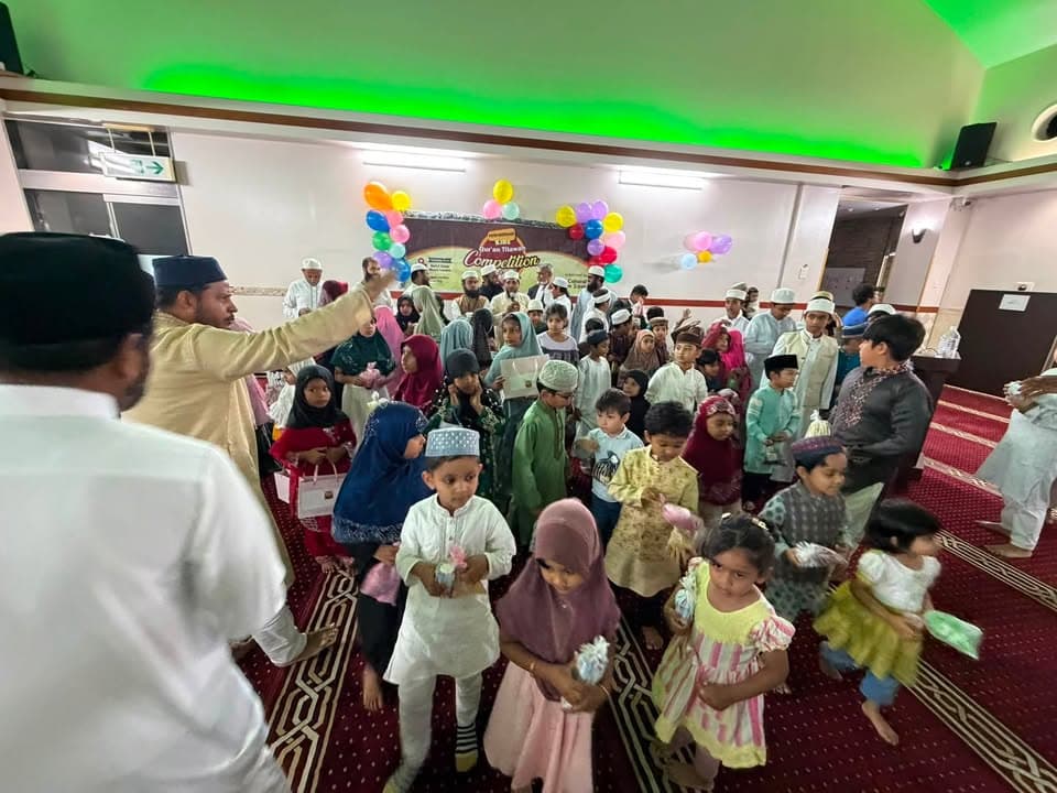 A large group of children in traditional Islamic attire, including boys wearing caps and girls in hijabs, gather in a mosque hall decorated with colorful balloons and a "Kids Qur'an Tilawat Competition" banner, while an adult man distributes small gifts to them.