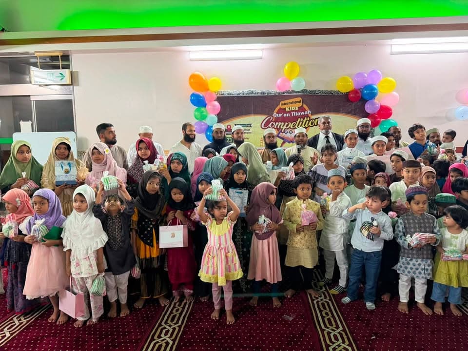 A large group of Muslim children and adult organizers pose together in a mosque hall decorated with colorful balloons and a banner reading "International Kids Qur'an Tilawah Competition," with many children holding gift bags.