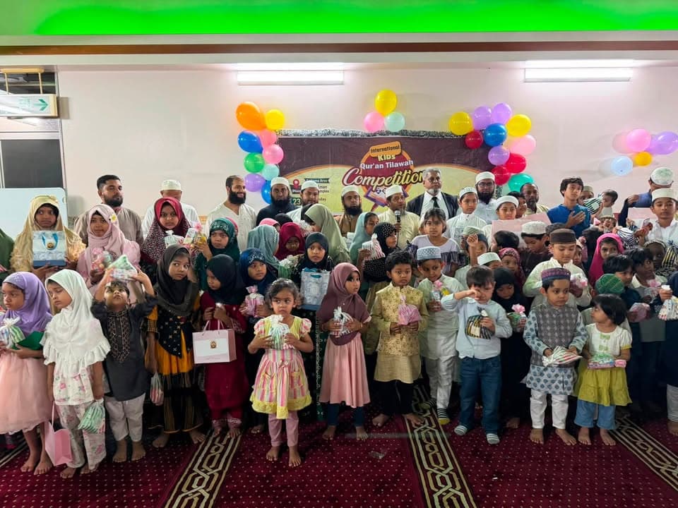 A large group of children in traditional dress, along with several adult organizers, stand together in a mosque hall decorated with colorful balloons and a banner reading "International Kids Qur'an Tilawah Competition," each child holding a gift bag.