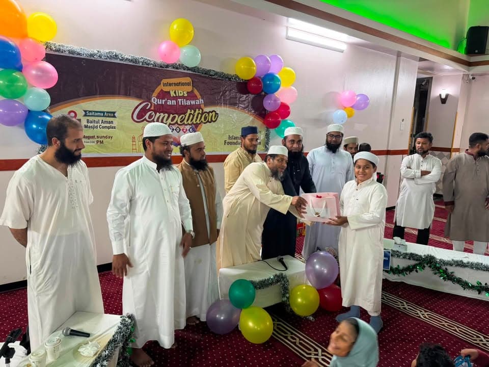 A boy in white robes receives a gift box from a bearded man in traditional dress on a stage decorated with colorful balloons and a banner reading "International Kids Qur'an Tilawah Competition," while several other men look on.