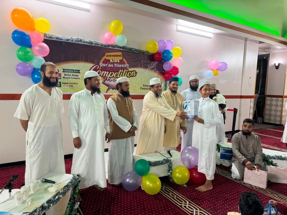 A boy in white robes receives an award from a bearded man in traditional dress on a stage decorated with colorful balloons and a banner reading "International Kids Qur'an Tilawah Competition," while other men in Islamic attire look on.