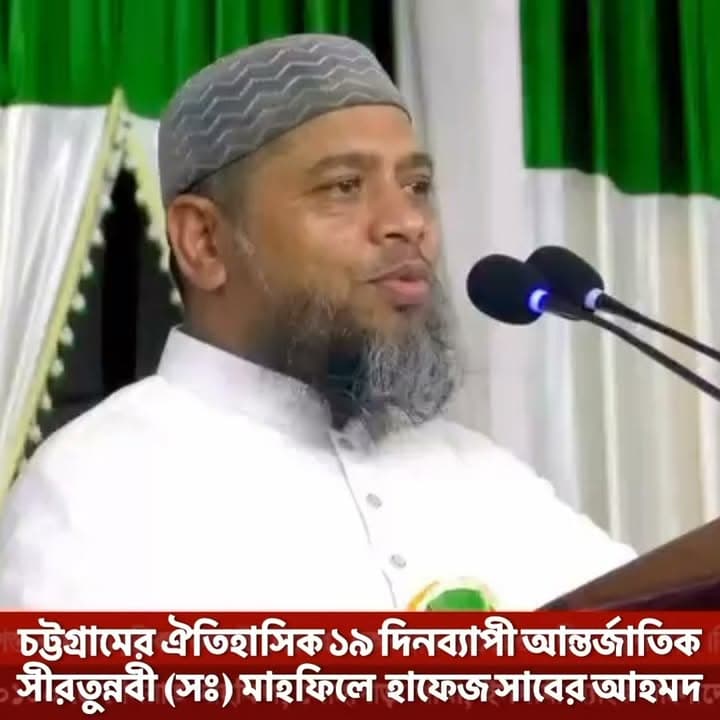 A bearded man wearing a gray cap and white kurta speaks into a pair of microphones at a podium, with a green and white curtain backdrop and Bengali text overlay at the bottom of the image.