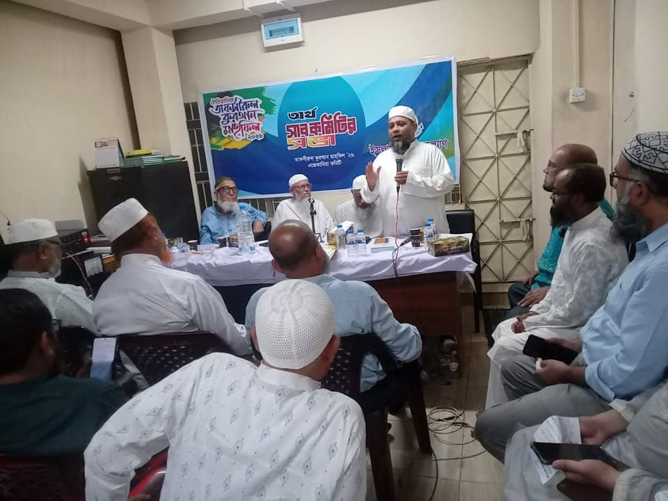 A man in a white robe and cap speaks into a microphone while seated at a head table with two other men in front of a blue banner with Bengali text, as a group of men in caps sit listening in a room.
