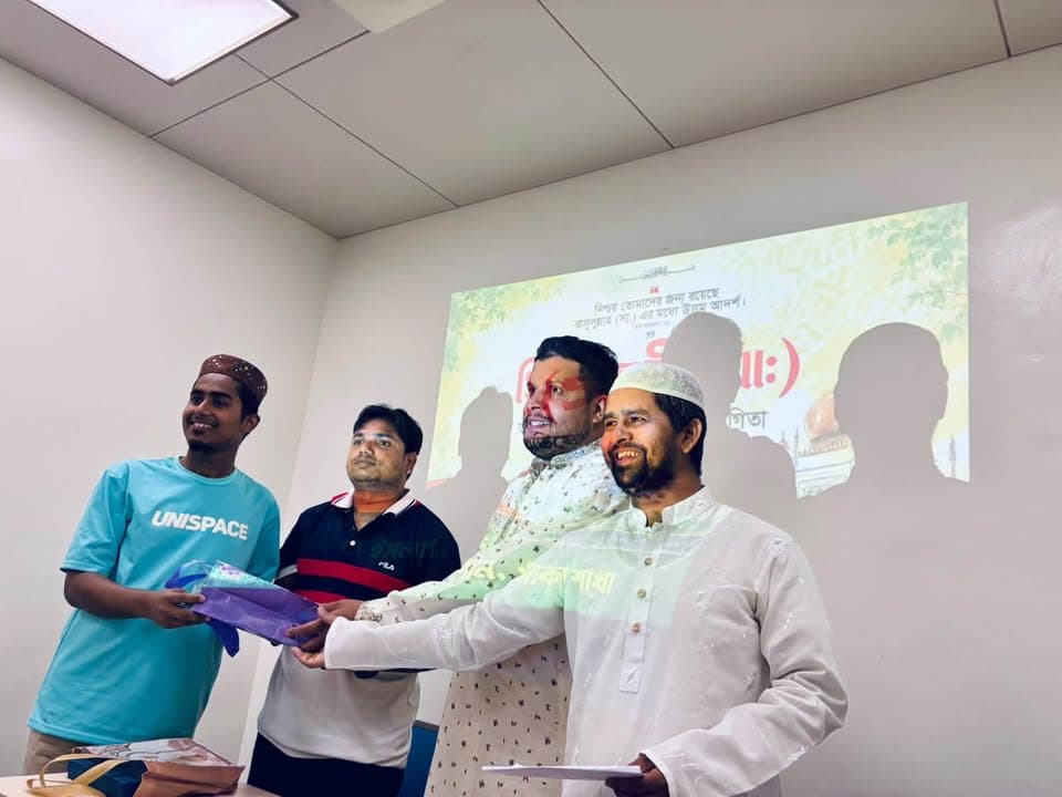 Four men stand together indoors handing over a wrapped gift, with a projected slide bearing Bengali text and a faded silhouette design displayed on the wall behind them.