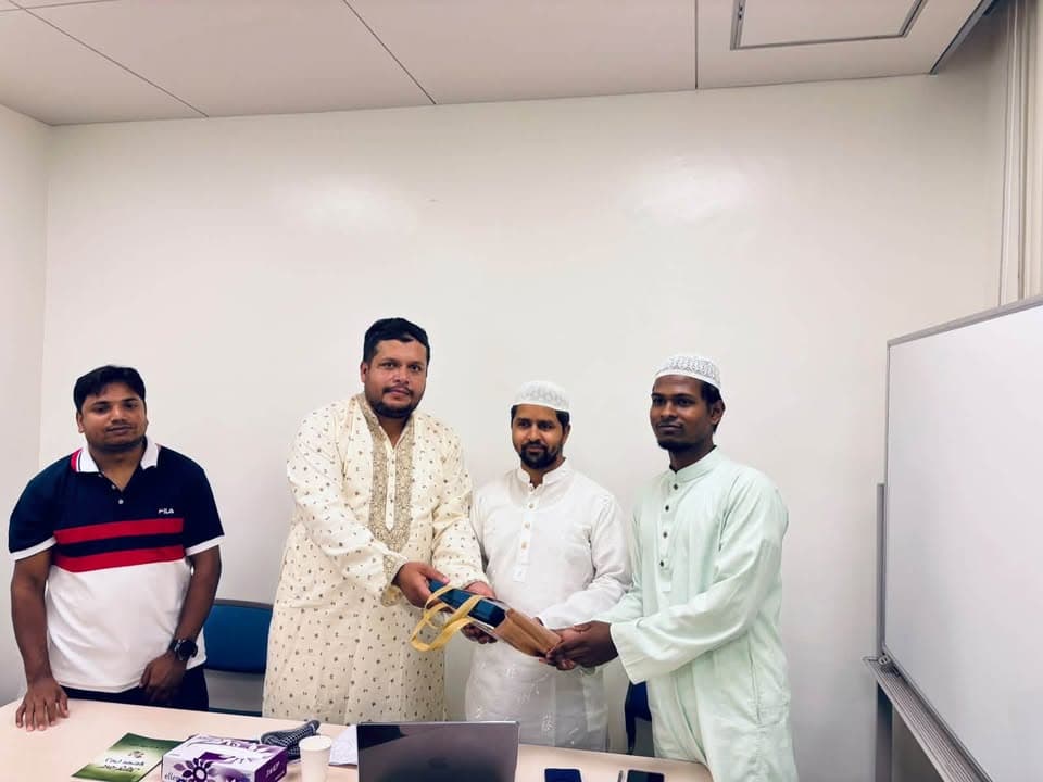 Four men stand in an indoor meeting room in front of a whiteboard; one man in traditional cream attire hands a rolled award or gift wrapped in blue ribbon to a younger man, while two other men look on.