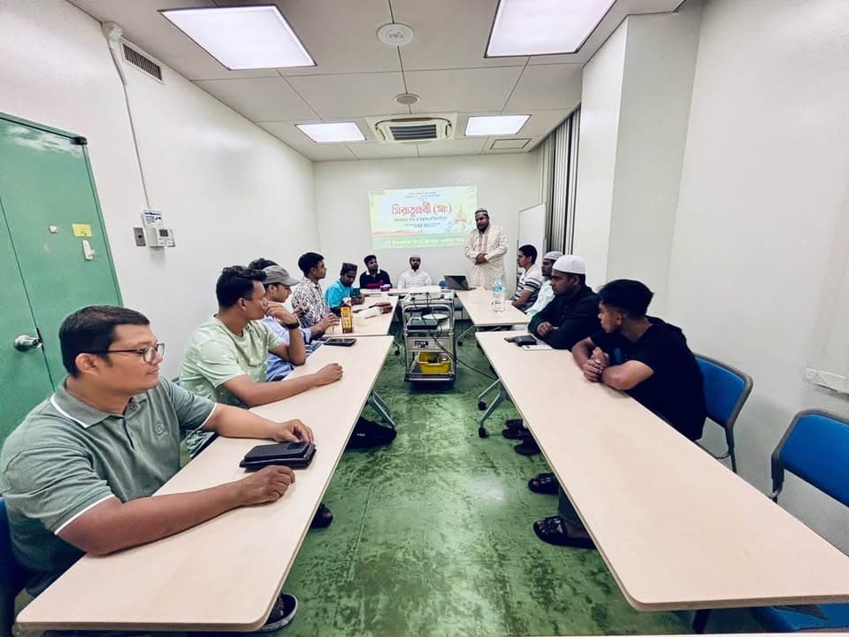 A group of men sit around tables arranged in a U-shape in a small conference room, listening to a speaker standing near a projector screen displaying text in Bengali.