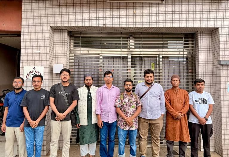 Nine men in casual and traditional clothing stand in a row outdoors, posing for a group photo in front of a building with a shuttered storefront and a Kyoto community center sign.