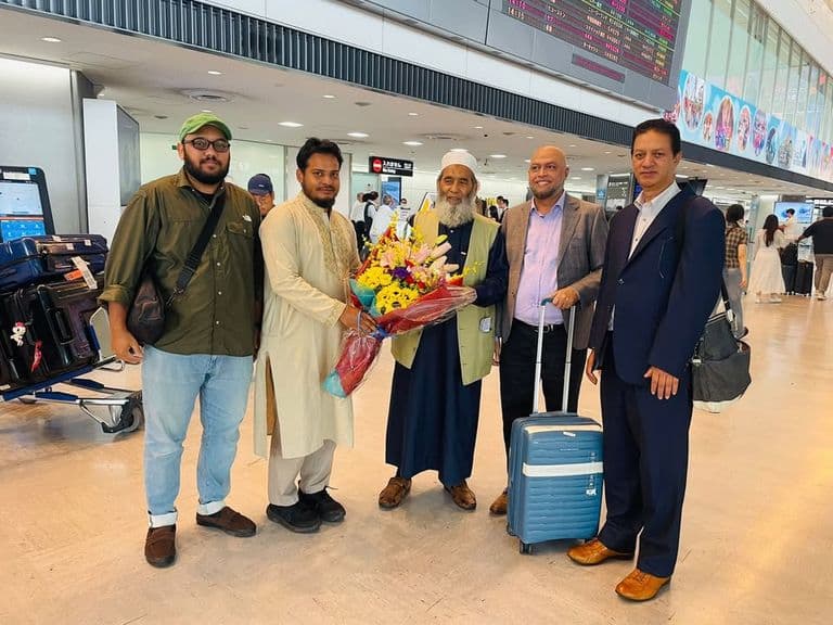 Five men stand together in an airport arrival hall, with an elderly bearded man in traditional dress holding a bouquet of flowers, a luggage cart and rolling suitcase beside them, and a flight information board in the background.