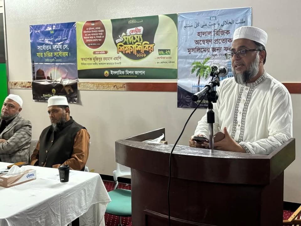A man in white traditional attire and a cap speaks at a wooden podium with a microphone, while two seated men listen behind a table, with Bengali and Arabic banners on the wall behind them.