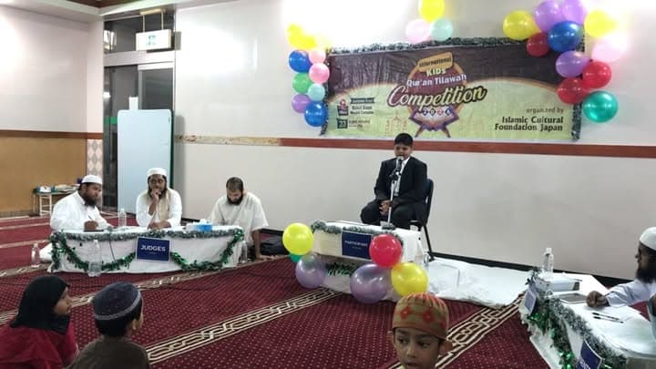A boy in a suit recites at a microphone on a stage decorated with balloons and a "International Kids Qur'an Tilawah Competition" banner, while a panel of judges sits at a table in front of him and other participants and audience members sit on the carpeted floor.