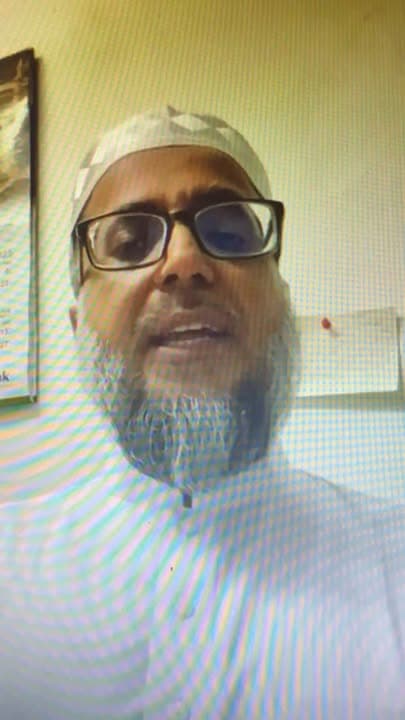A close-up screen capture of a man wearing glasses, a white prayer cap and a white robe, speaking during a video call, with a moiré pattern typical of a photographed screen.