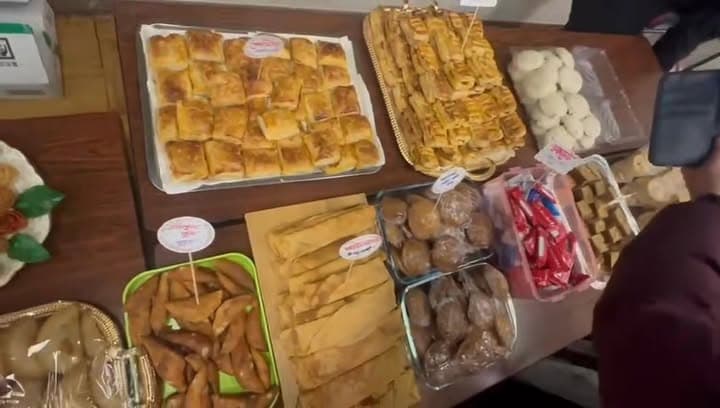 A table displays an assortment of traditional pastries and sweets, including baked pitha, samosa-like triangles, coconut balls, and wrapped candies, with price tags on small cards, as a person browses at the edge of the table.