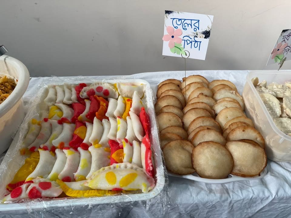 Trays of traditional Bangladeshi pitha desserts on a table, including colorful dumpling-shaped sweets and fried golden pithas, each tray labeled with a handwritten Bengali name card.