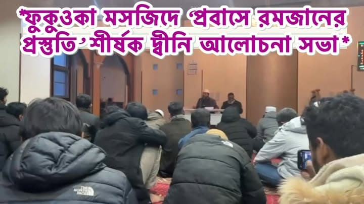 A group of men in winter coats sit on the carpeted floor of Fukuoka Masjid, listening to two speakers seated at a table at the front of the prayer hall.