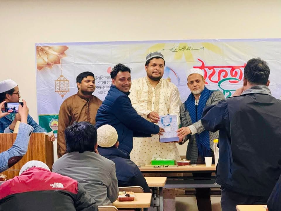 Men in traditional attire stand on a stage in front of a Ramadan-themed banner, with one presenting a gift plaque to another while attendees seated at tables watch and someone takes a photo.