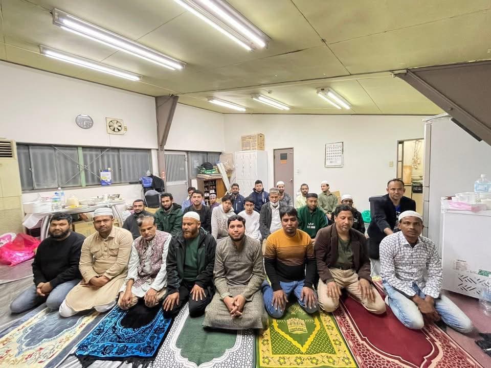 A large group of men kneel together on prayer rugs inside a community hall, posing for a group photo after gathering for a meal.