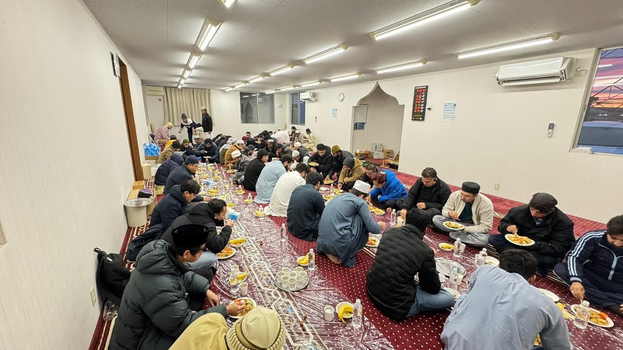 A long row of men sit cross-legged on a red patterned carpet along the walls of a mosque hall, eating from plates of food arranged on a plastic sheet, with prayer time boards and a mihrab visible at the back of the room.