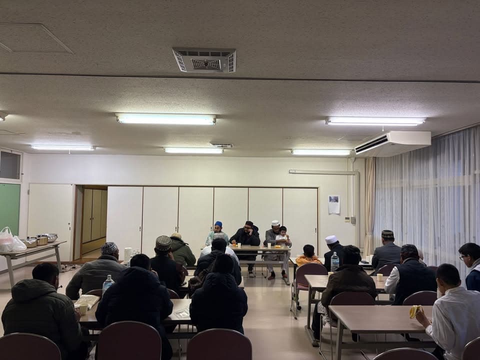 A group of men and children, many wearing prayer caps, sit at rows of tables in a community hall, listening as a few men speak at the front near a table set with food.