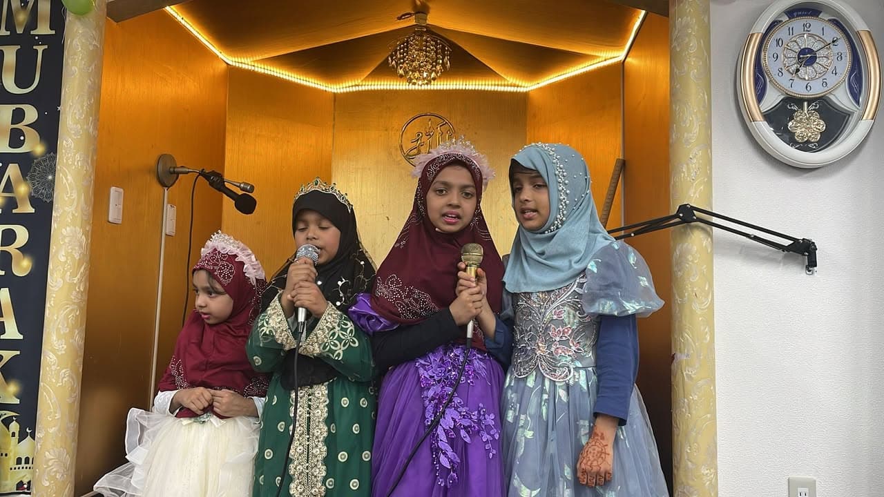 Four young girls in colorful hijabs and decorated dresses stand in front of a mosque's mihrab, two of them holding microphones as they perform.