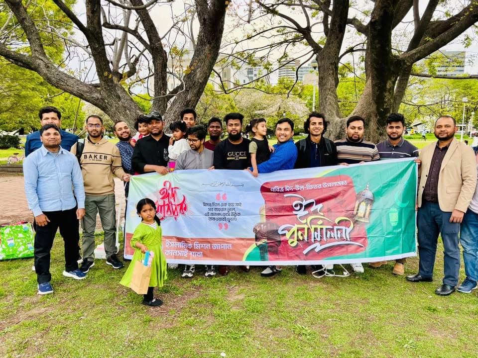 A group of men and children pose together under trees in a park, holding a large banner with Bengali text celebrating Eid, with a young girl standing in front of the banner.