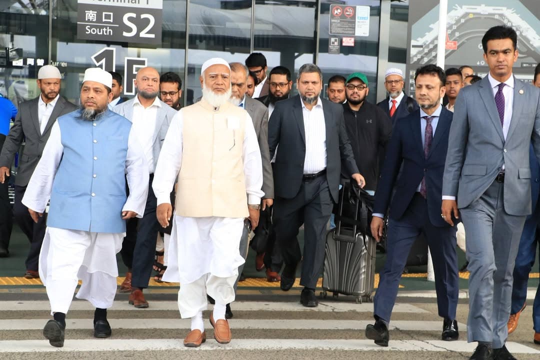 A group of men in suits and traditional white attire walk together outside the South exit S2 of Terminal 1 at an airport, with a man pulling a rolling suitcase among them.