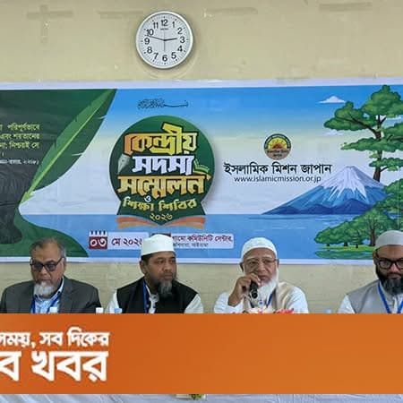 Four men in formal attire sit at a panel table on a stage, with one speaking into a microphone, in front of a large backdrop banner featuring Islamic Mission Japan's logo and Bengali text, with a wall clock mounted above.