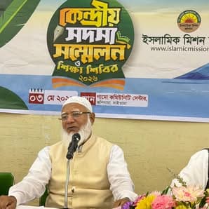 A man in a white cap and beard speaks into a microphone while seated at a table with a floral arrangement, in front of a stage backdrop with Bengali text and the Islamic Mission Japan logo.