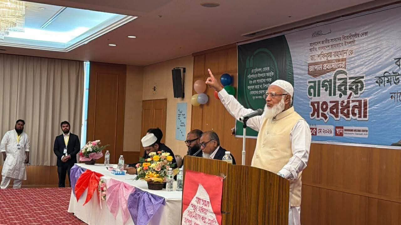 An elderly man with a white beard, wearing a cream-colored vest over a white kurta and a white cap, speaks passionately at a podium with a raised hand while gesturing with his index finger, in front of a green banner with Bengali text reading "নাগরিক সংবর্ধনা" (Civic Reception); several men are seated at a flower-decorated head table beside him, and two men stand near the wall at the back of the room.