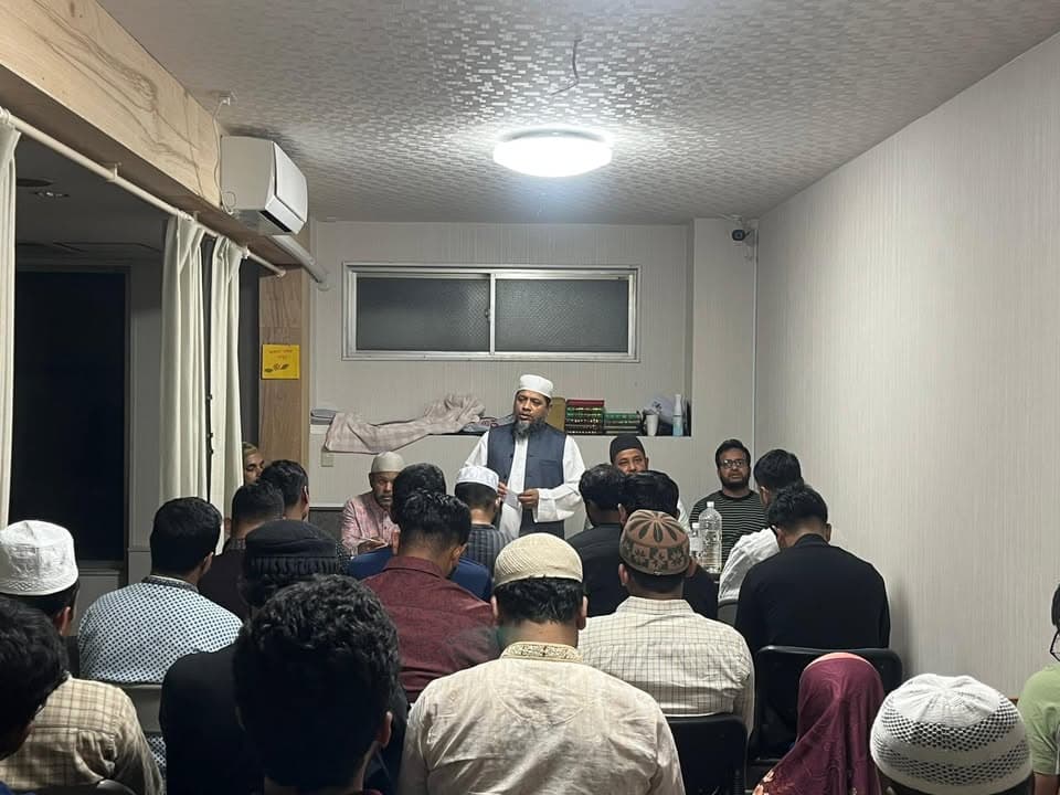 A man in a white cap and shirt stands and speaks while holding papers in front of a seated audience of men wearing caps, gathered closely together in a small indoor room.