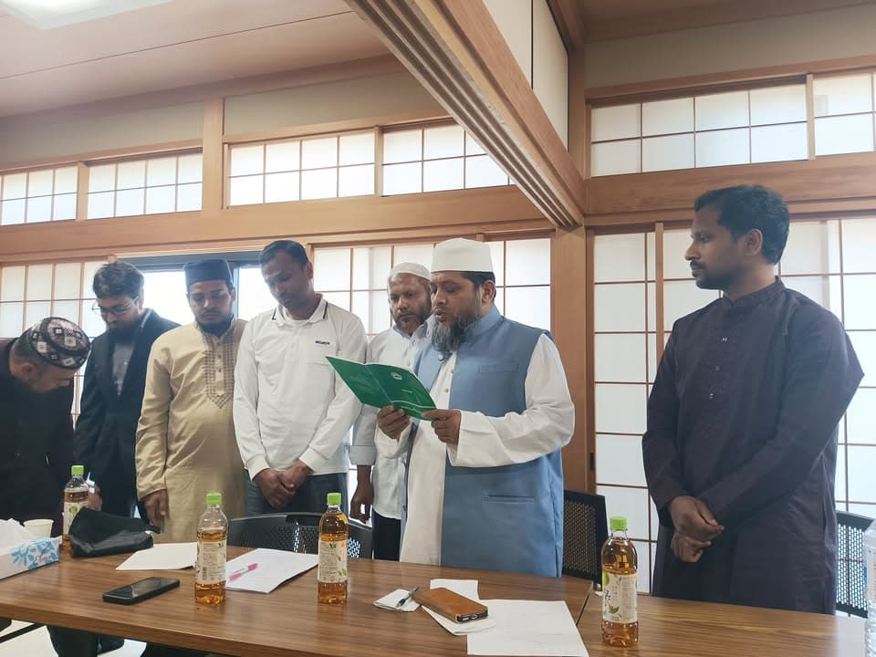 A group of men in traditional Islamic attire stand around a wooden table in a tatami-style room with shoji screens, while one man wearing a white cap and blue vest reads from a green booklet.