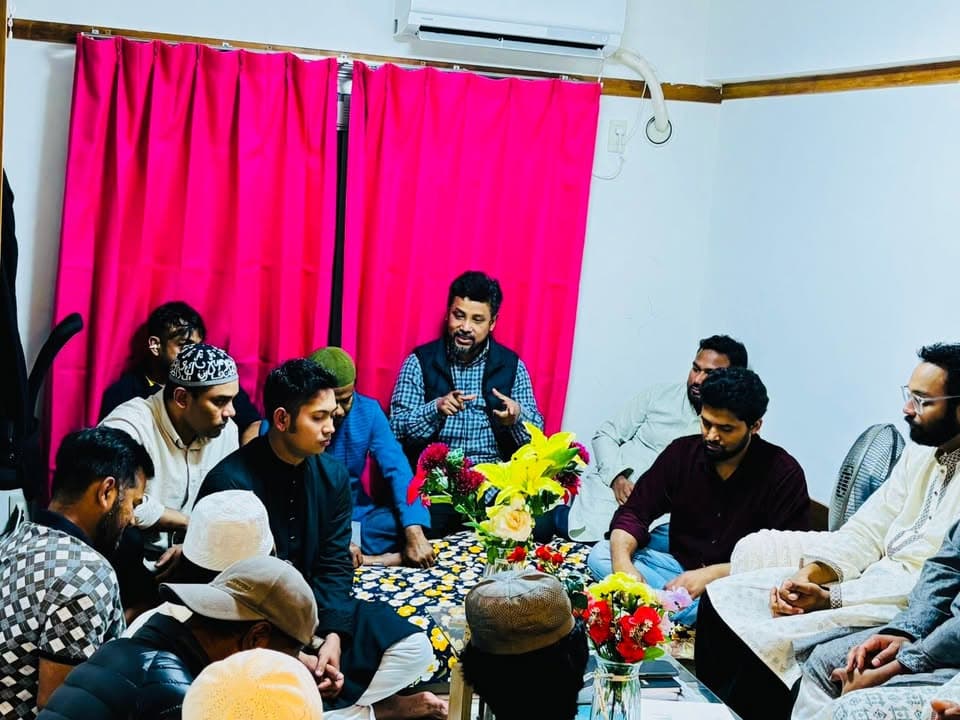 A group of men sit together on the floor around a low table decorated with flower vases, in a room with a pink curtain, as one man in the center speaks while gesturing with his hands.