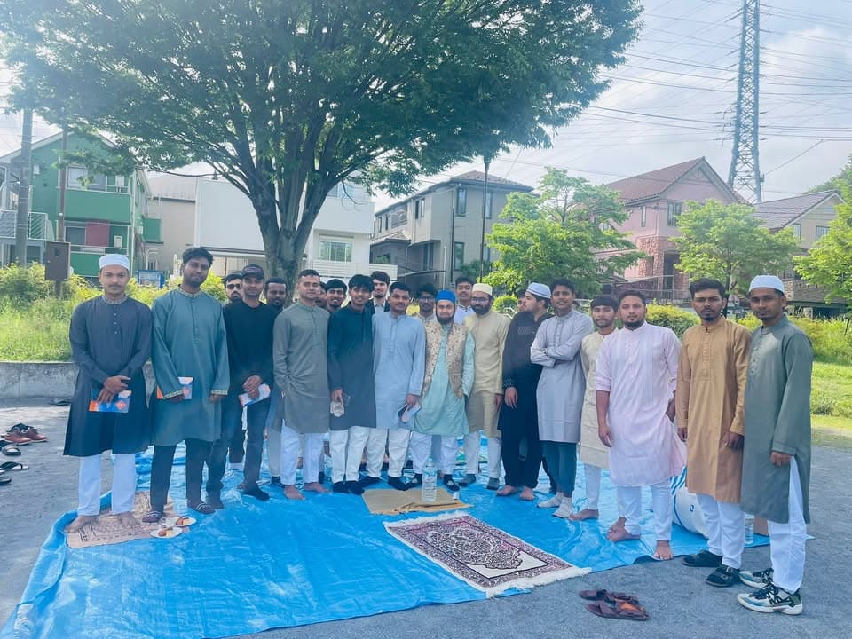 A group of young Muslim men in traditional attire stand together on blue tarps and prayer mats laid out under a tree in a park, with houses and a utility tower visible in the background.
