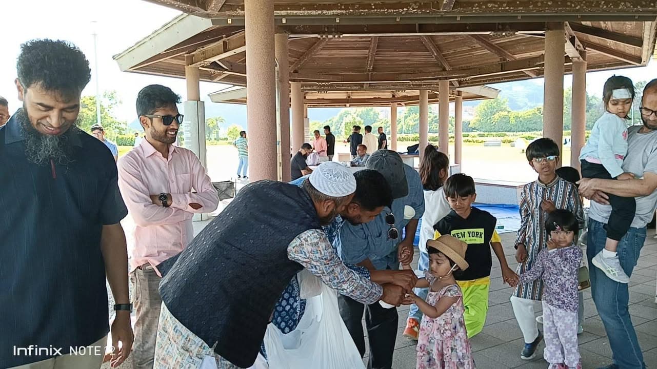 Under a large outdoor pavilion, men, women, and children gather as a man in a prayer cap bends down to greet a young child, with more families standing and holding hands nearby.