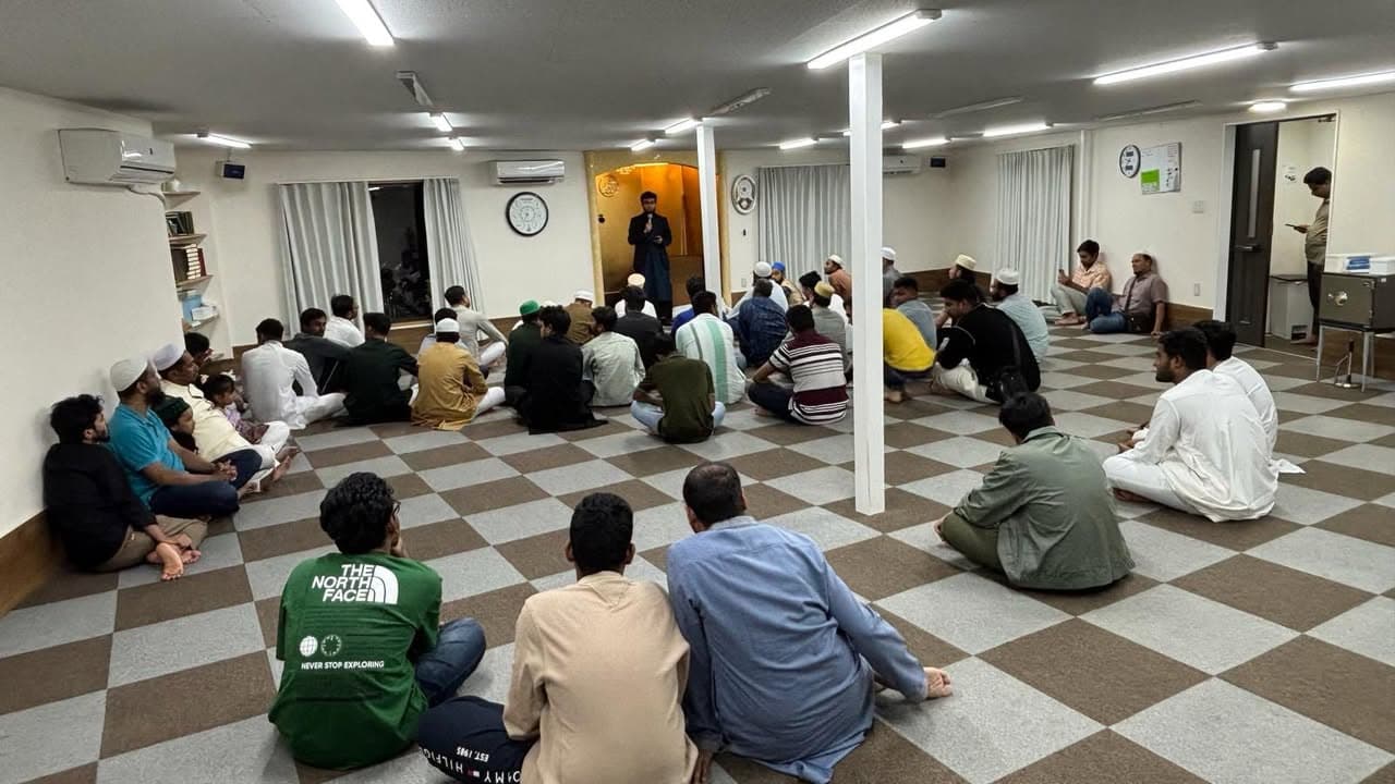 A large group of men and boys sit cross-legged on a checkered carpeted floor inside a mosque hall, listening to a man standing near the entrance speaking into a microphone.