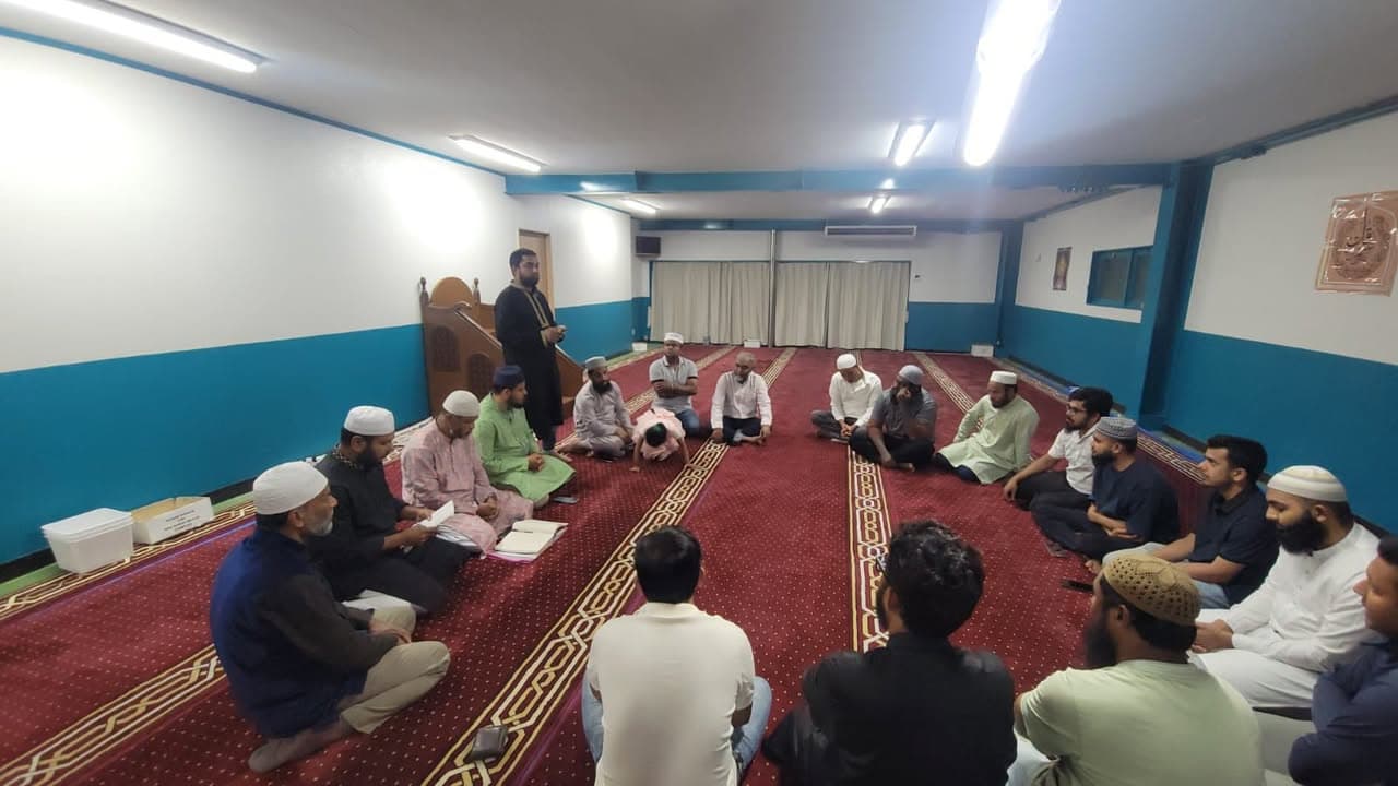 A group of men sit in a circle on the red patterned carpet of a mosque prayer hall, listening to a man standing near the mihrab who is speaking to them.