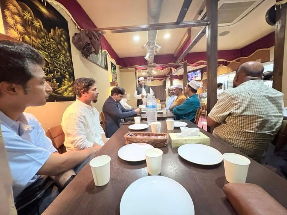 A group of men sit around a long table set with empty plates and paper cups in a restaurant with red-and-gold decor, listening to a man in a cap and vest standing at the head of the table.