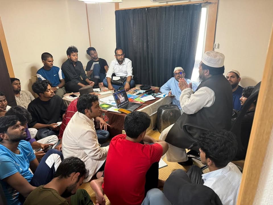 A group of men, mostly young students, sit closely together on a tatami-floored room, listening to an elder in a white robe and cap who gestures while speaking, with a laptop and papers on a low table in front of him.