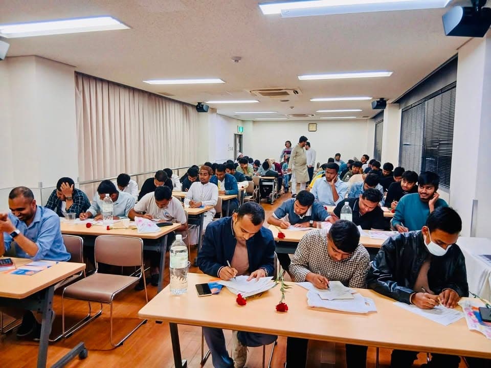 Rows of men sit at classroom tables writing on paper handouts, with water bottles and red carnations placed on the desks, in a community center hall.