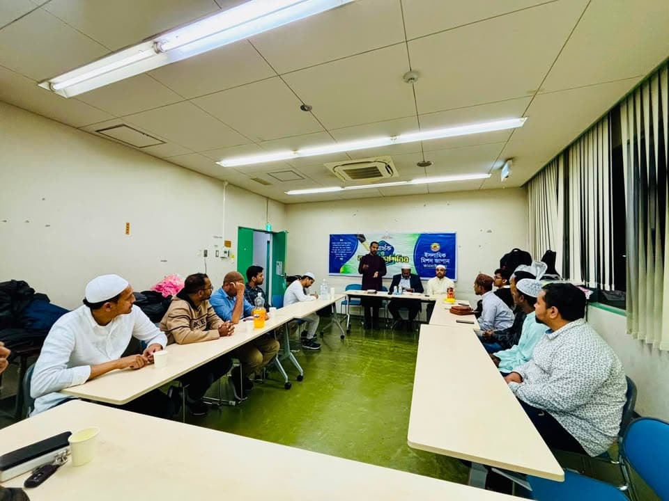Men seated around U-shaped rows of tables in a meeting room listen to a speaker standing at the front near a banner with Bengali text, with several attendees wearing white caps.