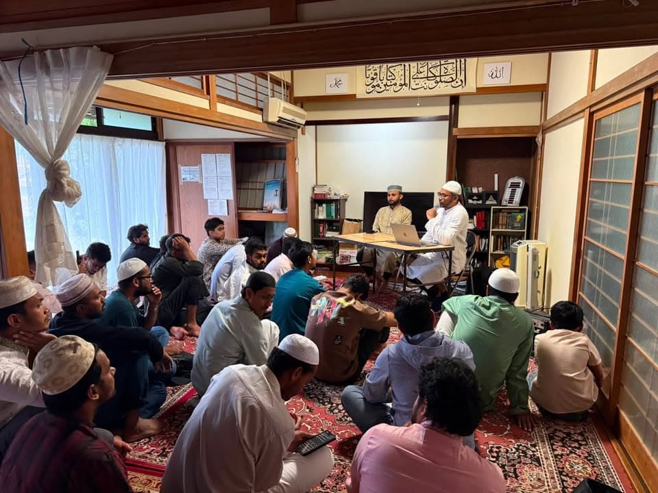 A group of men in traditional clothing and prayer caps sit on a carpeted tatami room floor, listening to two speakers seated at a table beneath Arabic calligraphy on the wall.