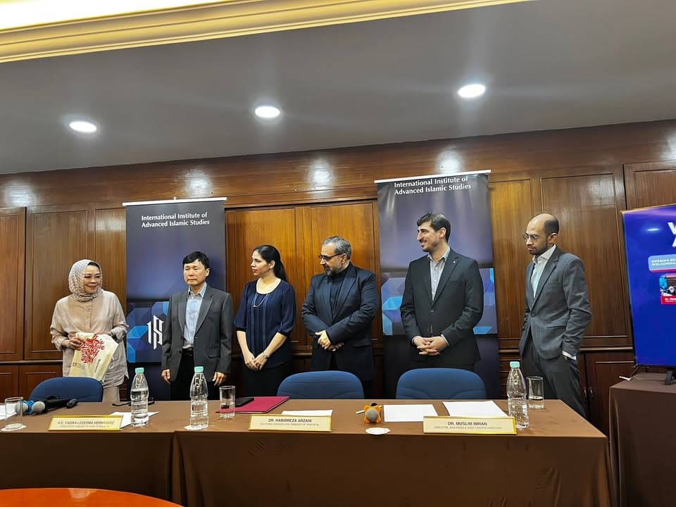 A group of six people, including men in suits and a woman in a headscarf, stand behind a conference table with name placards, in front of two "International Institute of Advanced Islamic Studies" banners.