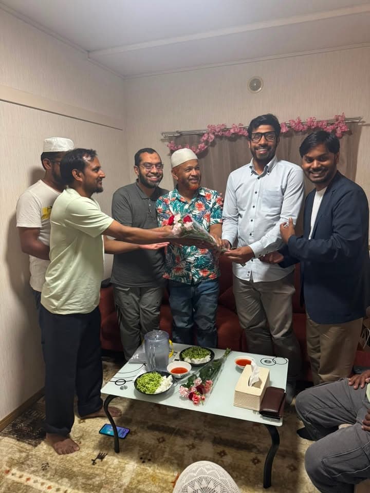 A group of men gather in a living room, with one man presenting a bouquet of flowers to another man wearing a floral shirt and white cap, while others stand around smiling; a low table with food dishes and flowers sits in front of a red sofa.