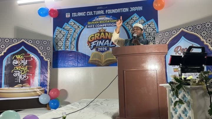 A man in a cap speaks passionately into a microphone at a podium, raising his hand, in front of a large banner reading "International Quran Recitation and Hifz Competition Grand Final 2026," decorated with balloons and mosque-shaped panels on either side.