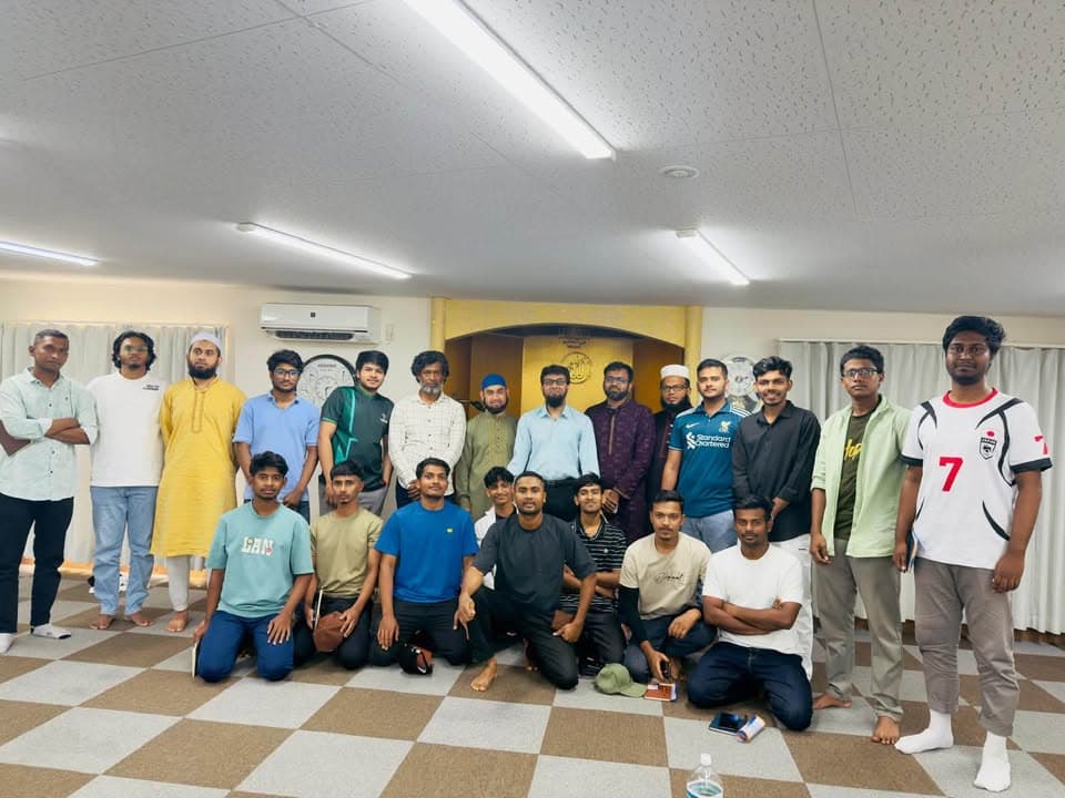 A large group of men, some standing and some kneeling, pose together for a group photo inside a mosque prayer hall with a mihrab visible in the background.