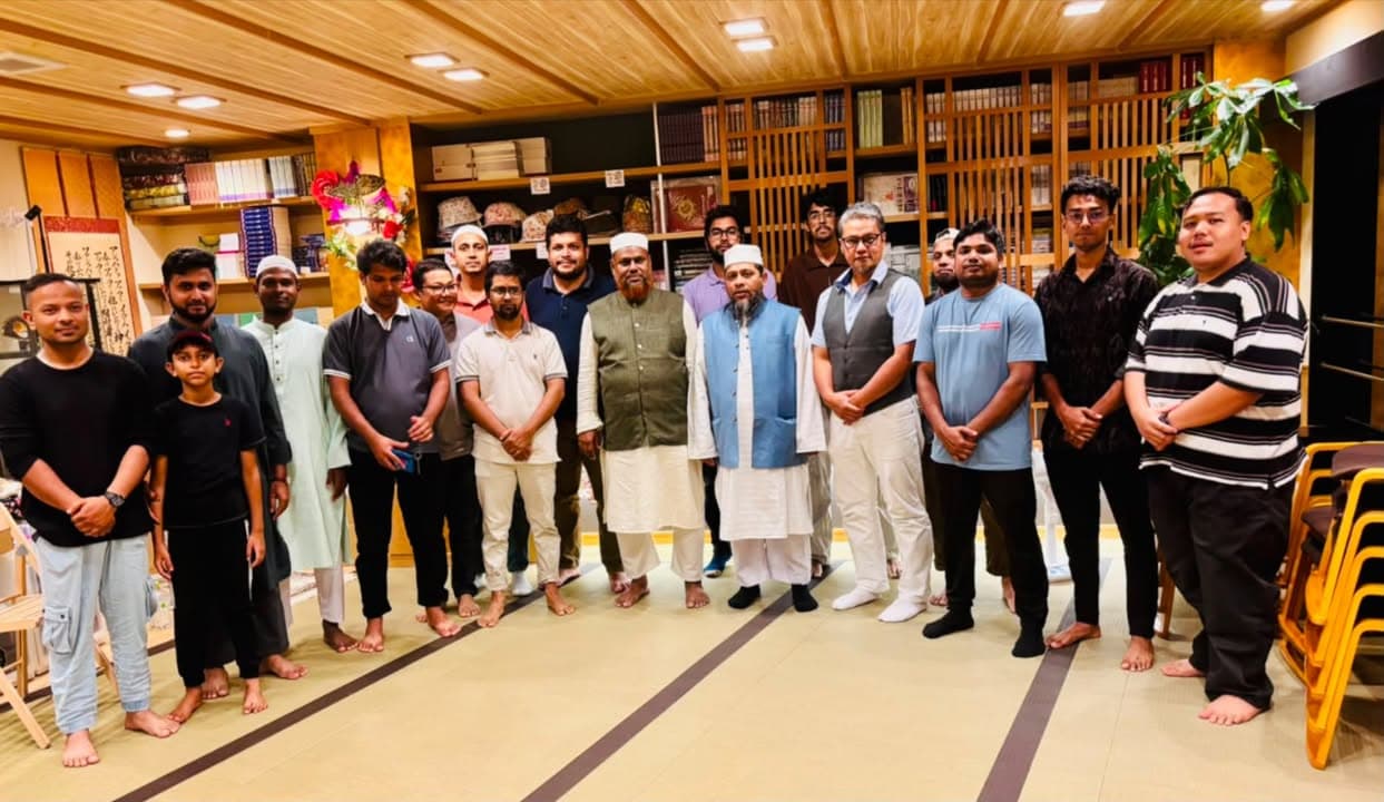A group of men and one boy, many wearing traditional Islamic attire and caps, stand in a line inside a wood-paneled room with bookshelves filled with religious texts in the background, posing together for a photo on tatami-style flooring.